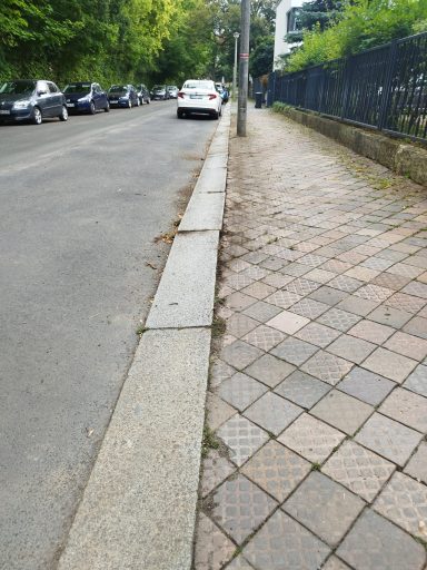 Straße mit geparkten Autos und schmalem Gehweg aus Pflastersteinen.