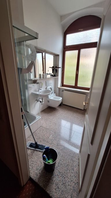 Sauberes Badezimmer mit Dusche, WC und großen Fenstern. Eimer und Putzutensilien sichtbar.