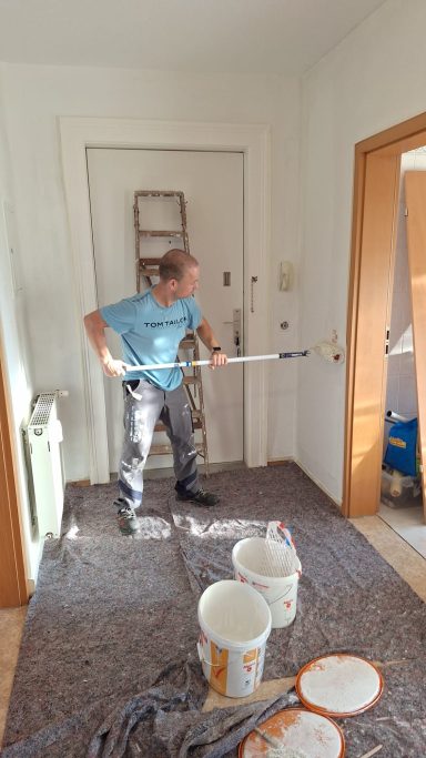 Mann streicht eine Wand in einem Raum mit Farbbehältern und Leiter im Hintergrund.