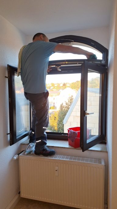 Mann reinigt ein Fenster in einem hellen Raum mit einem Eimer neben sich.